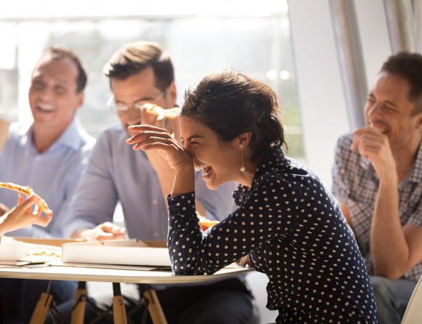 Indian woman laughing at funny joke eating pizza with diverse coworkers in office, friendly work team enjoying positive emotions and lunch together, happy colleagues staff group having fun at break